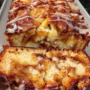 May include: A golden-brown apple cinnamon loaf cake with a moist crumb, featuring visible chunks of baked apple, a dark cinnamon swirl throughout the interior, and a decorative white sugar glaze drizzled over the top surface of the bread.