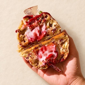 Puede incluir: Una galleta partida por la mitad con un exterior horneado de color marrón dorado, trozos de bayas rojas visibles y un relleno central blanco cremoso. La galleta tiene una apariencia texturizada y quebradiza con un glaseado rojo brillante y un pequeño adorno triangular transparente.