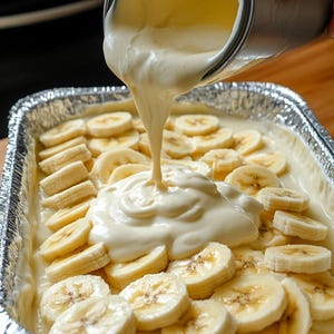 Op de afbeelding: Een rechthoekige aluminiumfolie pan gevuld met gesneden bananen en een romige, lichtgekleurde saus die uit een metalen container wordt gegoten. De bananen zijn in een enkele laag gerangschikt en de saus is dik en glad.