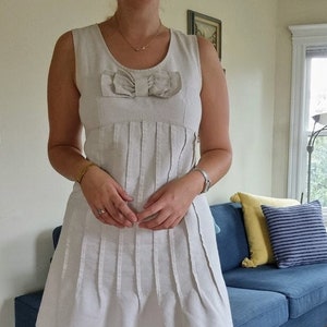 May include: A white sleeveless dress with a bow detail at the neckline. The dress has a pleated skirt and is made of a lightweight fabric.