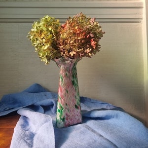 Puede incluir: Un jarrón de vidrio colorido y con forma de espiral con una boca ancha contiene un ramo de hortensias secas en tonos de verde, rosa y naranja. El jarrón está sobre un paño azul.