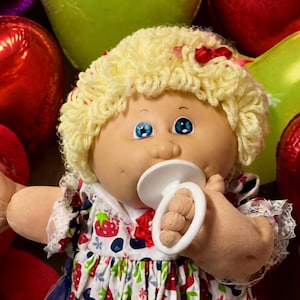 May include: A doll with blonde yarn hair and blue eyes wearing a white dress with a red strawberry and blue blueberry print, white lace-trimmed socks, and white shoes, holding a white pacifier, surrounded by teddy bears and heart balloons.