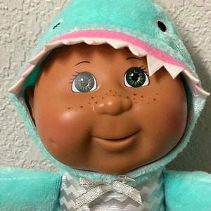 Peut inclure: Une poupée en peluche portant un costume de requin bleu et blanc. La poupée a la peau brune, les yeux verts et un grand sourire. La poupée est assise sur une couverture colorée.