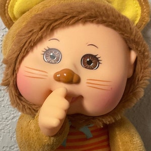 May include: A brown plush toy wearing a brown and yellow lion costume. The toy has a brown nose, brown eyes, and a pink blush on its cheeks. The toy is standing on a colourful patterned blanket.