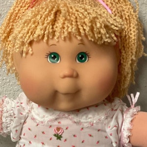 Può includere: Una bambola Cabbage Patch Kid con capelli biondi in lana, un fiocco rosa e un vestito bianco con fiori rosa. La bambola indossa calzini bianchi con pizzo e sta in piedi su un patchwork colorato.