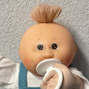 May include: A vintage Cabbage Patch Kid doll with a peach-colored face, brown eyes, and a tuft of light brown hair. The doll wears a white shirt, blue overalls, and white sneakers with blue stripes. It holds a pacifier and a blue bag.