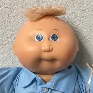 May include: A Cabbage Patch Kid doll wearing a blue shirt, blue jeans, and a belt with a cartoon animal print. The doll has blonde hair and blue eyes. The doll is holding a small pink mesh bag.