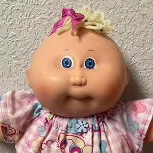 Può includere: Una bambola Cabbage Patch Kids con un top a quadri rosa e bianco e pantaloncini bianchi con finiture in pizzo. La bambola ha gli occhi blu, un fiocco rosa e la scritta "Bubbles" sulla camicia.