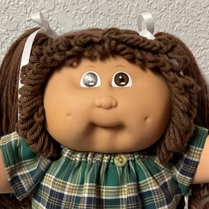 May include: A vintage doll with brown yarn hair styled in pigtails, wearing a green and blue plaid outfit. The doll has a tan face with painted eyes and a smiling expression. It is wearing white lace shoes.
