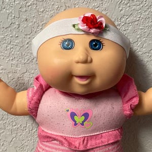 Peut inclure: Poupée Cabbage Patch Kids vêtue d'une tenue rose avec un motif de papillon. La poupée a les yeux bleus, un bandeau blanc avec une fleur rouge et des chaussettes roses. Les bras de la poupée sont tendus.