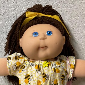 May include: A Cabbage Patch Kid doll with brown hair, blue eyes, and a yellow bow. The doll wears a yellow top and shorts with a bee pattern, and white lace-trimmed socks and shoes. A pink bag is visible.