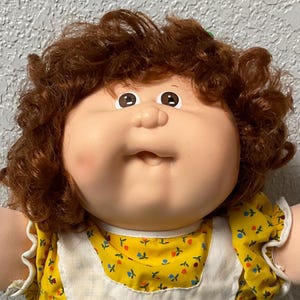 Peut inclure: Une poupée Cabbage Patch Kid aux cheveux bruns bouclés, portant une robe jaune avec un tablier vert et une bordure en dentelle blanche, tenant un sac rose.