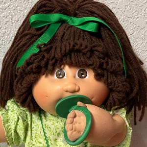 Peut inclure: Une poupée vintage avec des cheveux bruns en fil, un ruban vert et une sucette verte. La poupée porte une robe verte à motifs floraux, des chaussettes blanches et des chaussures à volants. Un petit sac avec un jouet est attaché.