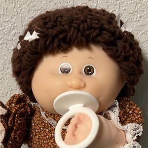 May include: A vintage doll with brown yarn hair, wearing a floral print dress with lace trim, white socks, and white shoes. The doll has a pacifier in its mouth and is holding it with its hands. A blue bag is next to the doll.