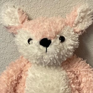 May include: A plush toy fox with pink and white fur. The fox has a white chest, pink paws, and a black nose. The ears are white with pink accents. The toy is sitting on a blue surface.