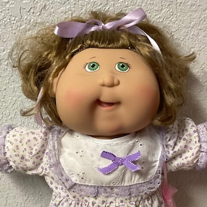 May include: A vintage doll with a round face, green eyes, and light brown hair. The doll wears a white dress with a purple floral pattern, a white bib with a purple bow, and white shoes with lace-trimmed socks. A purple ribbon adorns the doll's hair.