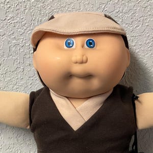 May include: A Cabbage Patch Kid doll wearing a brown vest, tan pants, and a matching cap. The doll has blue eyes and brown shoes with white laces. A black accessory hangs from the doll's arm.