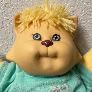 Op de afbeelding: Een gele pluche kat met blond garenhaar die een lichtblauw T-shirt draagt met een klein kattenmotief. De kat staat op een gestreepte stof.