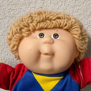 May include: A vintage Cabbage Patch Kid doll with blonde yarn hair, wearing a red, blue, and yellow shirt, teal shorts, and white trainers. The doll has a smiling expression and outstretched arms.