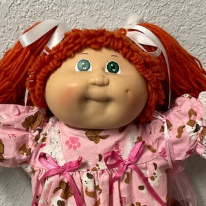 May include: A vintage doll with red yarn hair in pigtails, white ribbons, and green eyes. The doll is dressed in a pink dress with a dog and paw print pattern, lace trim, and pink bows. White lace-trimmed socks and white shoes complete the outfit.