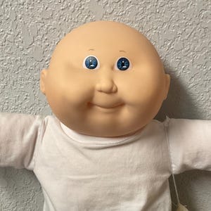 May include: A Cabbage Patch Kids doll with a bald head, blue eyes, and a smiling expression. The doll is wearing a white t-shirt, dark blue denim shorts, and white shoes. A small white bag hangs from the doll's side.