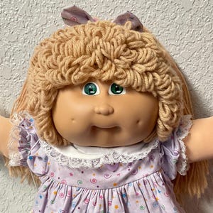 May include: A vintage Cabbage Patch Kid doll with blonde yarn hair and green eyes. The doll wears a lavender dress with a star and swirl pattern, a white lace collar, and matching socks with lace trim. The doll's arms are outstretched.
