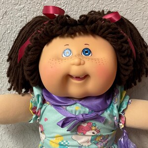 May include: A vintage doll with brown yarn hair styled in pigtails, adorned with red ribbons. The doll has blue eyes, freckles, and a smiling expression. It wears a light blue dress with a colorful print, a purple collar, and white shoes. A purple bag is held in its hand.
