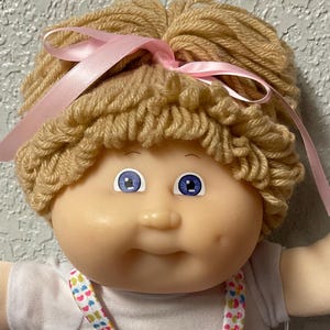 May include: A Cabbage Patch Kid doll with blonde yarn hair, blue eyes, and a pink bow. The doll is wearing a white shirt, a pink and white dress with a heart pattern, and pink socks with white ruffles.