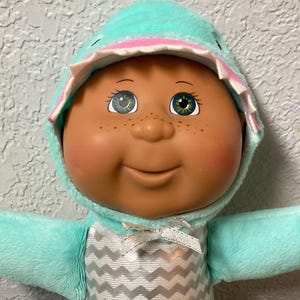 May include: A vintage Cabbage Patch Kids doll wearing a turquoise and white chevron patterned outfit with a matching hood. The doll has brown skin, freckles, and green eyes. The doll is sitting on a colorful quilt.