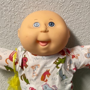 Puede incluir: Una muñeca Cabbage Patch Kid con una cara color melocotón, ojos azules y una expresión sonriente. La muñeca lleva una camisa blanca de manga larga con gráficos de animales coloridos y pantalones verdes. Una bolsa amarilla está unida a la muñeca.