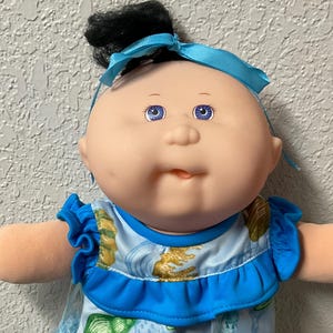 May include: A vintage Cabbage Patch Kid doll with a blue bow, black hair, and blue eyes. The doll is dressed in a blue dress with seashell patterns, white lace socks, and blue shoes. The doll's arms are outstretched.