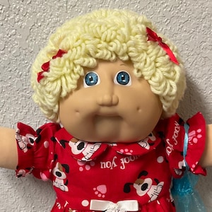 Puede incluir: Muñeca Cabbage Patch Kid con pelo rubio de lana y ojos azules. La muñeca lleva un vestido rojo con estampado de perro y lazo blanco. Zapatos y calcetines negros. Una pequeña bolsa azul con un pompón rosa está adjunta.