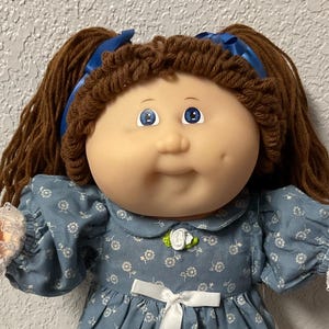 May include: A vintage doll with brown yarn pigtails, blue eyes, and a rosy complexion. The doll wears a blue floral dress with white lace trim, white socks, and white shoes. A white bow adorns the dress. The doll is posed on a colorful quilt.