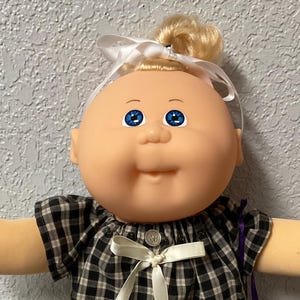 May include: A Cabbage Patch Kid doll with blonde hair in a bun, blue eyes, and a round face. The doll is dressed in a black and white checked top, grey trousers, white shoes with lace trim, and a white bow. A purple bag is visible.