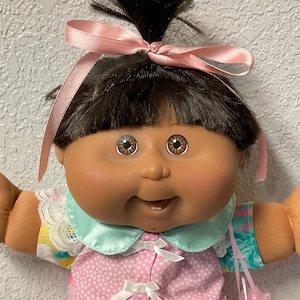 May include: A vintage Cabbage Patch Kids doll with dark hair styled in a topknot with a pink bow. The doll wears a pink and white dress with a ruffled skirt, pink socks, and pink shoes. The doll has brown eyes and is holding a pink bag.