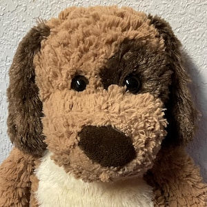 May include: A plush dog toy with brown and tan fur. The dog has dark brown ears, a dark brown nose, and a white patch on its chest. The toy has black eyes and is sitting upright with its legs extended.
