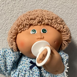 Puede incluir: Muñeca Cabbage Patch Kid vintage con un vestido azul floral con ribete de encaje blanco y zapatos a juego. La muñeca tiene cabello de hilo marrón claro, ojos verdes y un chupete. Un pequeño cubo de Rubik cuelga de una bolsa de malla rosa.