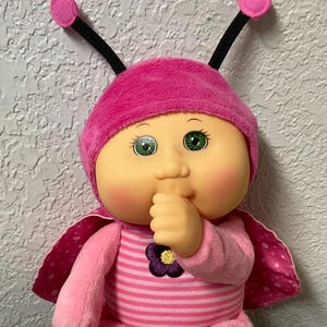Op de afbeelding: Een pluche pop in een roze en wit gestreept outfit, met een bijpassende roze hoed en vleugels. De pop heeft groene ogen en een vinger in de mond. De pop zit op een kleurrijke quilt.