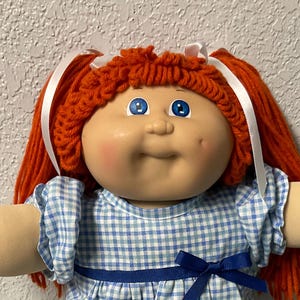 May include: A vintage Cabbage Patch Kids doll with red yarn hair, blue eyes, and rosy cheeks. The doll is dressed in a blue and white gingham dress with a blue bow and white lace-trimmed socks. A birth certificate and adoption papers are visible.