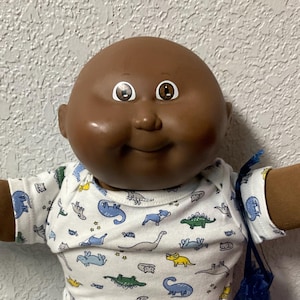 Peut inclure: Une poupée Cabbage Patch Kid vintage à la peau foncée, vêtue d'une chemise blanche à motifs de dinosaures, d'un pantalon bleu clair et de chaussures blanches. La poupée a les yeux bruns et une expression souriante. Elle tient un sac bleu.