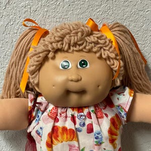 May include: A vintage Cabbage Patch Kid doll with blonde yarn hair tied with orange ribbons. The doll wears a white outfit patterned with croissants, cupcakes, and berries. The doll's shoes are white with a lace overlay.