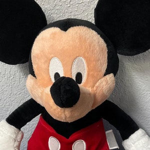 Pode incluir: Um grande brinquedo de pel&uacute;cia do Mickey Mouse com orelhas pretas, uma camisa vermelha e branca e sapatos amarelos. O brinquedo est&aacute; sentado em uma superf&iacute;cie azul.