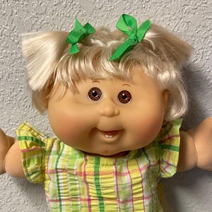 Peut inclure: Une poupée Cabbage Patch Kid vintage avec des cheveux blonds en couettes, ornées de nœuds verts. La poupée porte une tenue à carreaux jaune et vert, des chaussures roses et des chaussettes en dentelle blanche. La poupée a un sourire et les bras tendus.