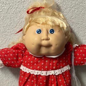Puede incluir: Muñeca Cabbage Patch Kid vintage con cabello rubio y ojos azules. La muñeca viste un vestido rojo con cuello blanco y estampado de fresas, calcetines y zapatos blancos. Una cinta roja está atada en el pelo de la muñeca. Objeto de colección.