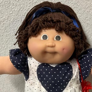 May include: A vintage doll with brown hair and a blue ribbon. The doll wears a white dress with navy blue polka dots and a navy blue heart-shaped design. The doll's dress has navy blue trim and short sleeves. The doll is wearing white shoes.