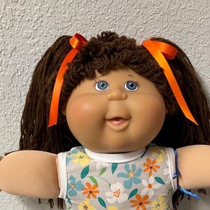 May include: A vintage doll with brown hair styled in pigtails, adorned with orange ribbons. The doll has blue eyes and is wearing a floral print dress with white shoes. The doll is a classic toy.