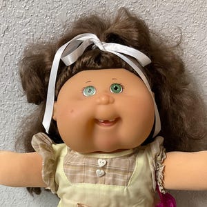 Peut inclure: Poupée vintage aux cheveux bruns, yeux verts et expression souriante. La poupée porte une robe jaune avec un corsage à carreaux, un nœud blanc et des chaussures blanches. Elle tient un sac rose. Un jouet classique.
