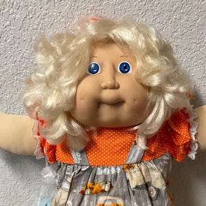 Peut inclure: Une poupée vintage aux cheveux blonds bouclés et aux yeux bleus, vêtue d'un haut orange à pois et d'une robe à bretelles grise avec un imprimé de camion de citrouilles. La poupée porte également des chaussures blanches à bordure en dentelle. Le thème général est l'automne.