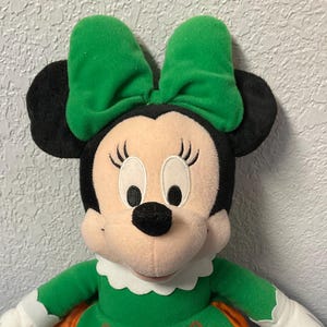 Puede incluir: Mu&ntilde;eca de peluche de Minnie Mouse con un vestido verde con detalles en naranja y un lazo verde a juego. La mu&ntilde;eca tiene orejas negras, nariz negra y guantes blancos. La mu&ntilde;eca est&aacute; sentada sobre una superficie azul.