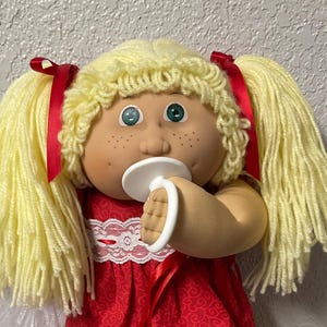 Peut inclure: Une poupée vintage avec des cheveux blonds en laine tressés en couettes, ornées de rubans rouges. La poupée porte une robe rouge avec un col en dentelle blanche et des chaussures blanches. Elle a les yeux verts, des taches de rousseur et une sucette. Un petit sac de perles est dans sa main.